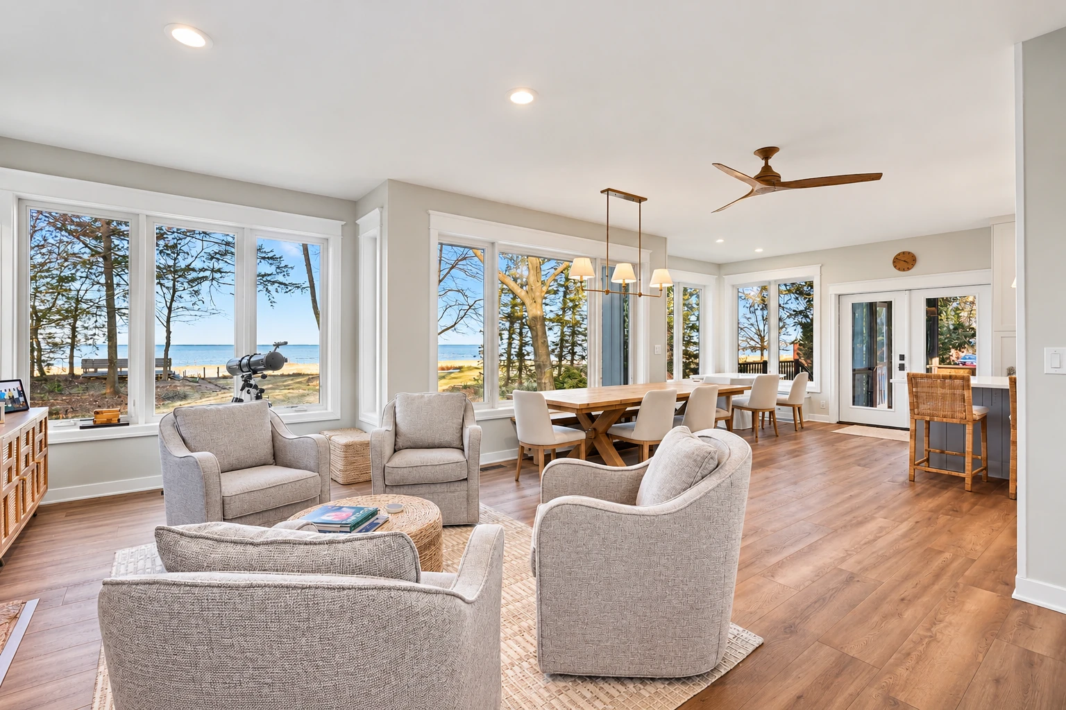 A bright four-season sunroom with broad windows, a sitting area, and dining table overlooks a wooded lakeshore.