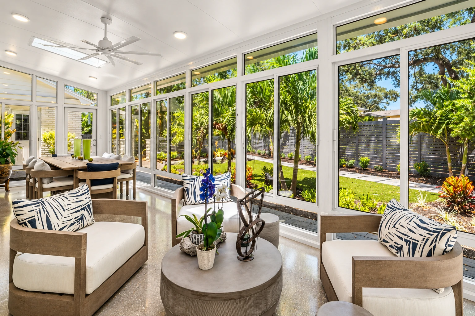 A bright four-season sunroom has white-framed glass walls, modern lounge seating, a dining table and views of a tropical garden.