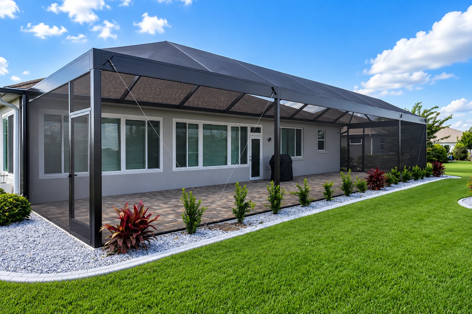 Large black-framed patio cover with mesh sides shades a paved area beside a gray house and green lawn.