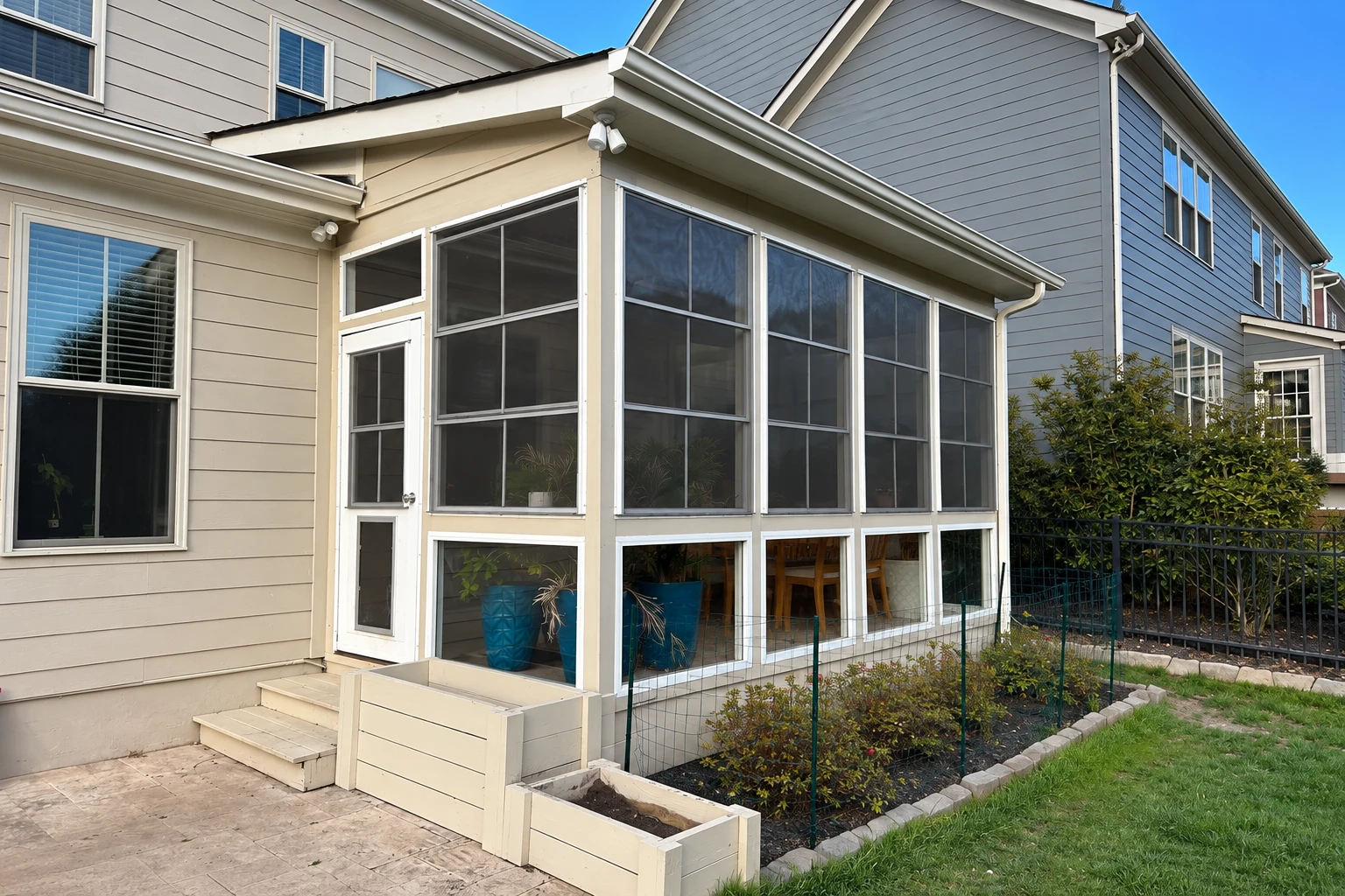 A patio enclosed with white-framed screens and windows, attached to a beige-sided house beside a small garden.