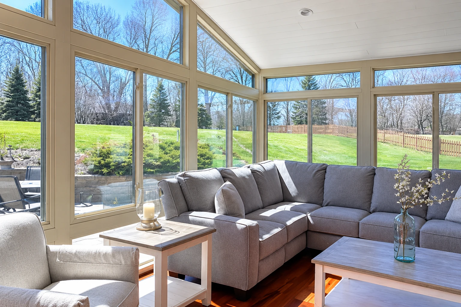 A converted patio sunroom with large windows, a gray sectional and light wood tables overlooking a lawn.