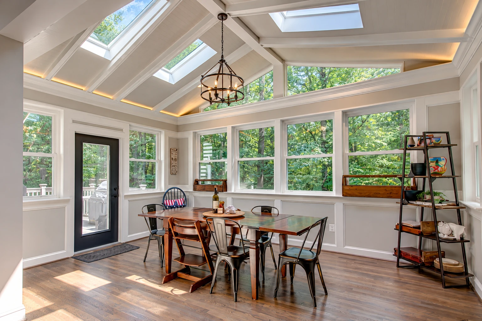 A sunroom dining area features a high vaulted ceiling with skylights, large windows, a dark entry door and a wooden table.