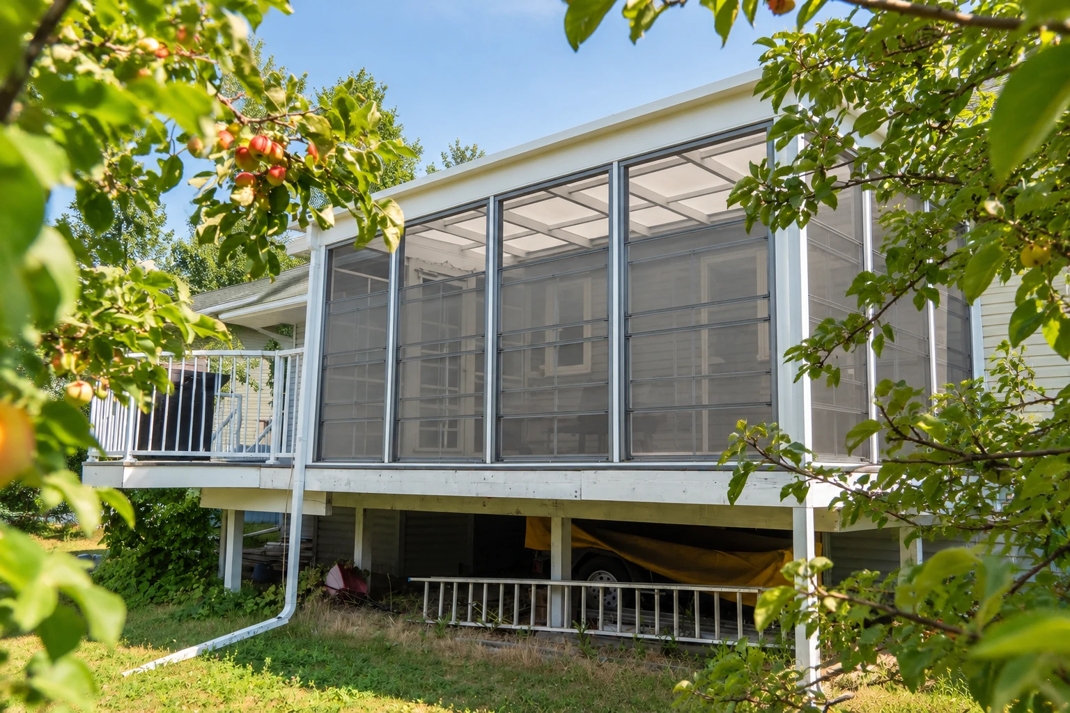 An elevated three-season screen room with white framing and dark mesh panels is partly framed by leafy branches.