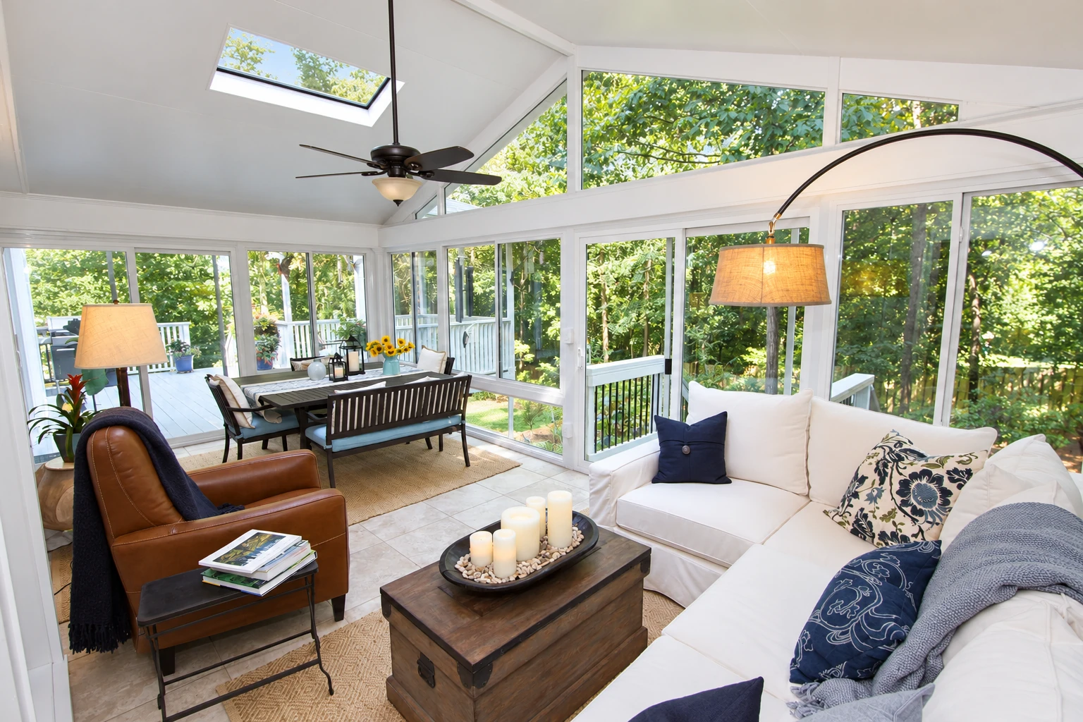 A bright vinyl sunroom with tall windows, a skylight, white sofa and a dining set overlooking leafy trees.